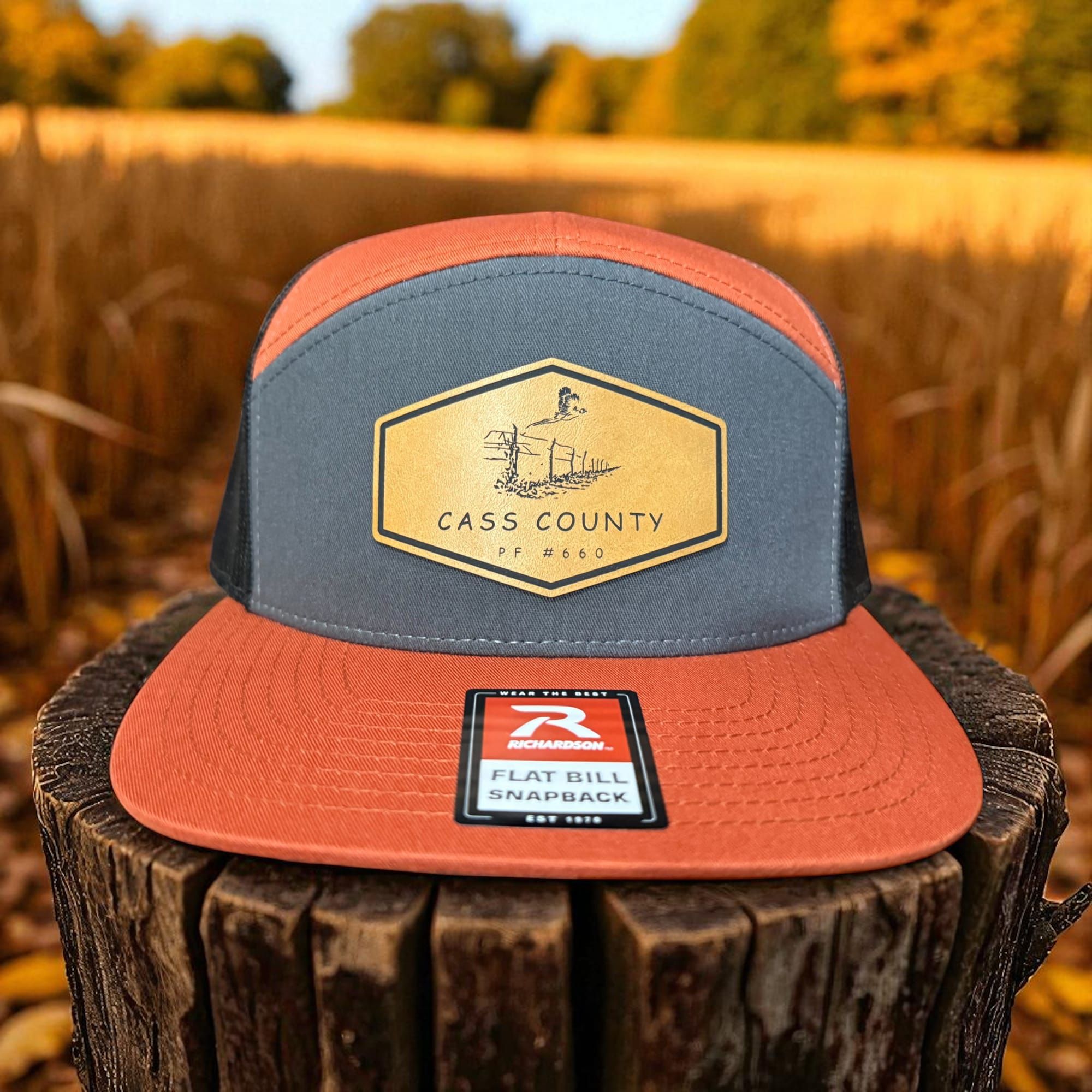 Orange and slate flat-bill snapback with a tan hexagon Cass County patch on a stump in a wheat field