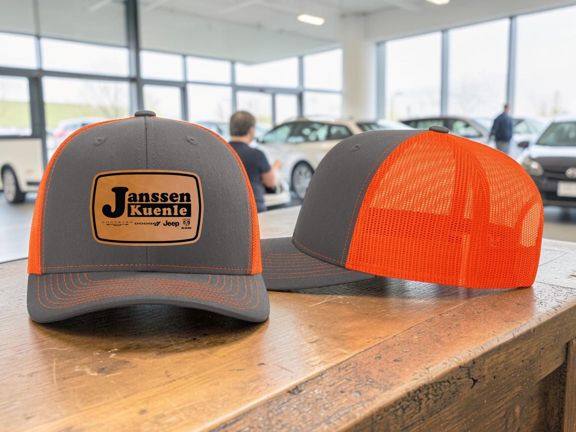 Two charcoal and neon orange trucker hats with engraved Janssen Kuenle leatherette patches in a car showroom