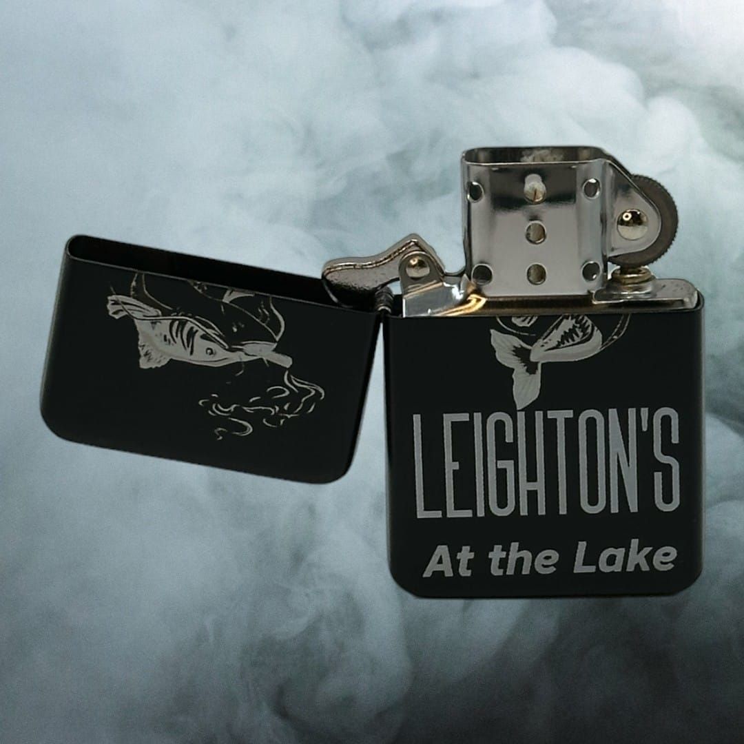 Black flip lighter engraved Leighton's at the Lake with a fish, lid open in smoke