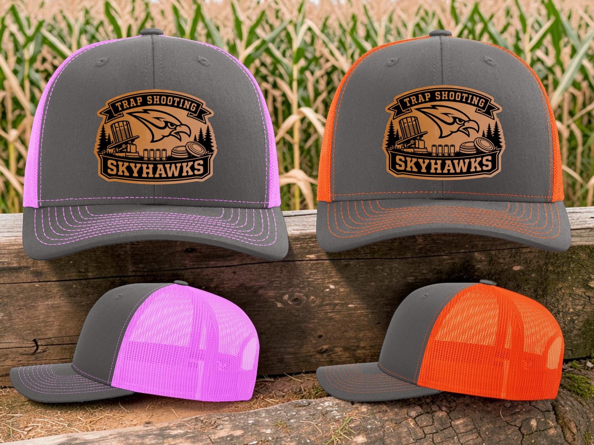 Charcoal trucker hats with pink and orange mesh and Skyhawks Trap Shooting leatherette patches in front of a cornfield